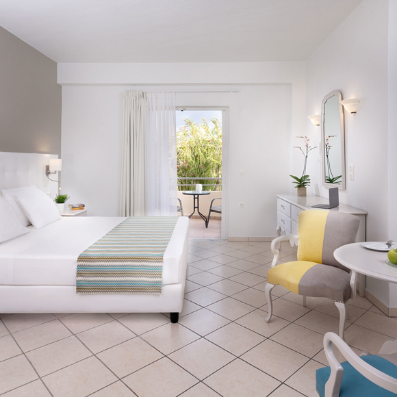 Double room with white furnishing, cozy seating area, wall-mounted TV and balcony view of greenery