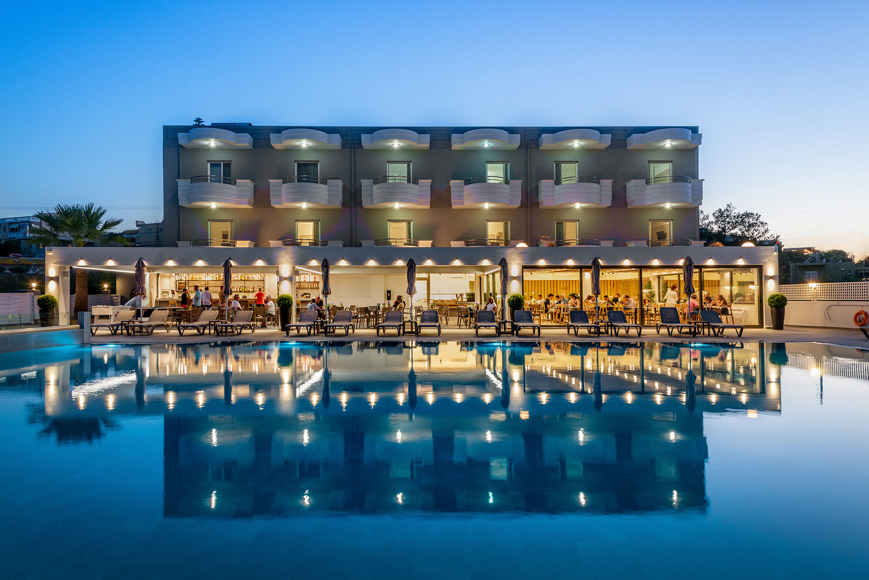 Hotel exterior with illuminated balconies and restaurant area reflected in the pool at dusk
