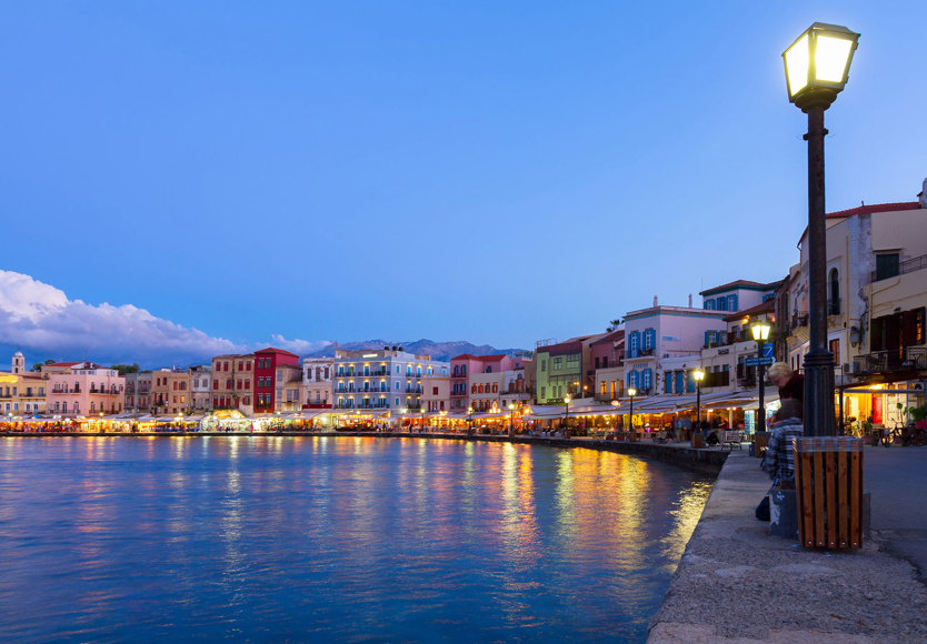 The waterfront road of Chania's Venetian harbor at dusk, lined with shops and restaurants